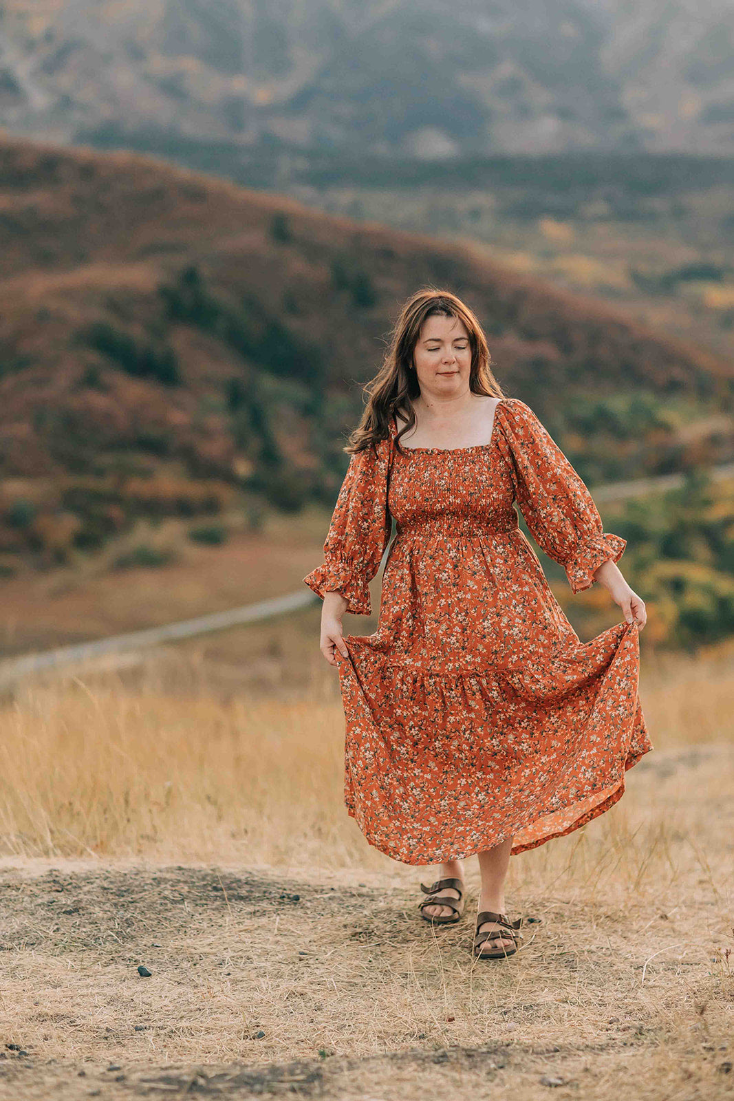 Woman in a rust-colored (orange) floral dress standing in a scenic outdoor setting with mountains.