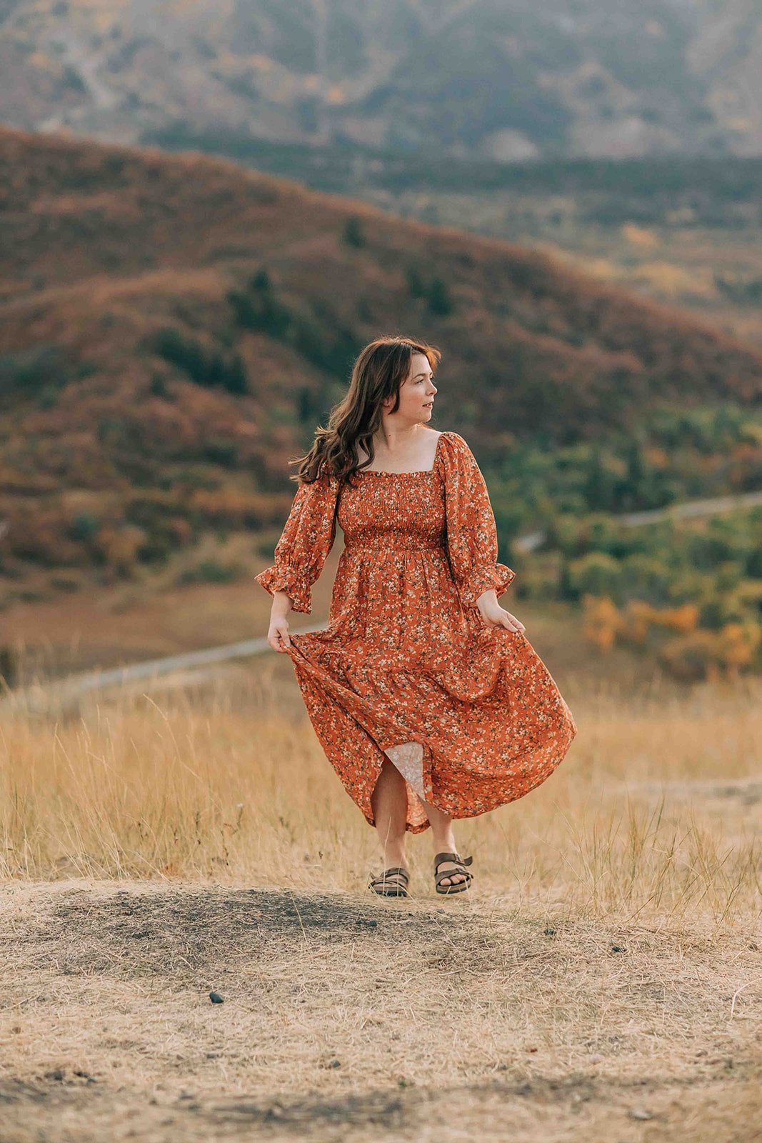 Woman in an orange floral smocked bodice, maxi dress walking on a dirt path with mountains in the background