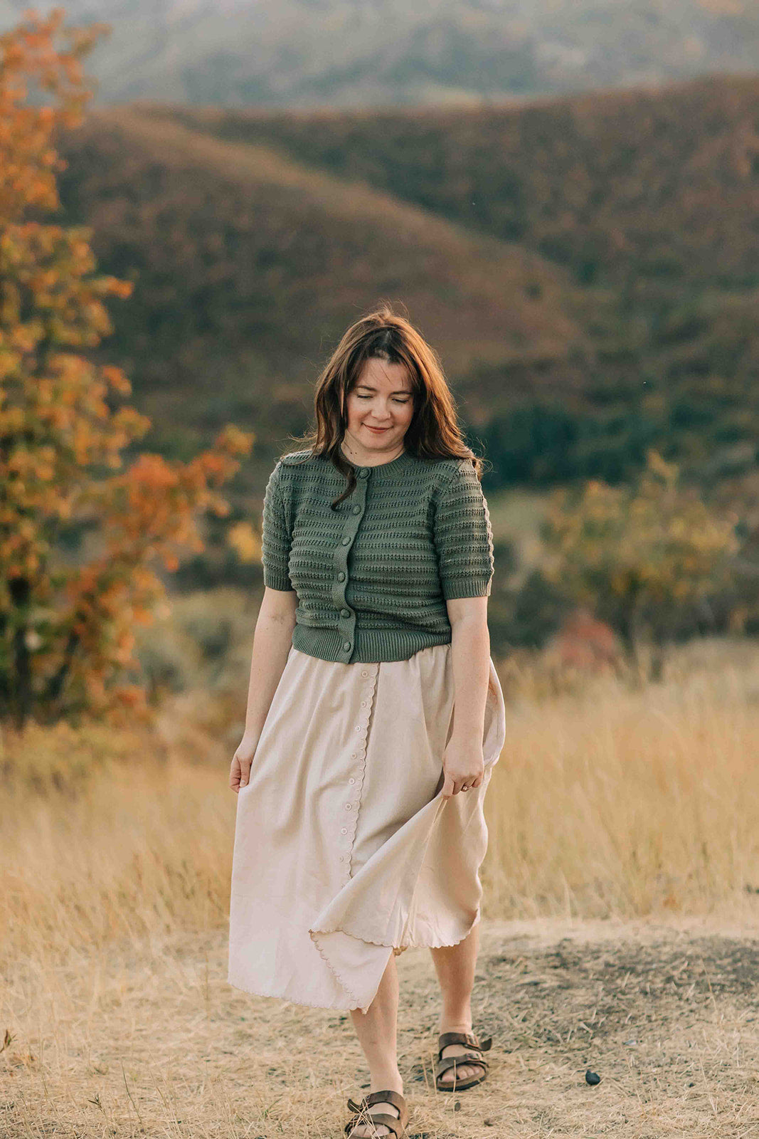 Woman modeling a green stripe textured half sleeve cardigan, and beige scalloped skirt, standing in a field with mountains in the background