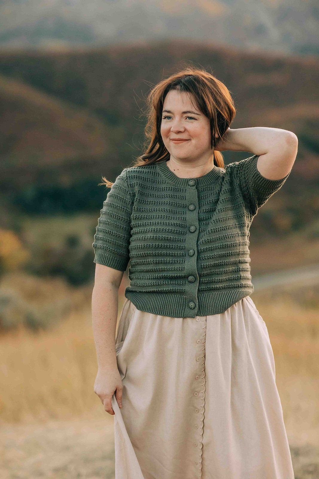 Woman wearing a green knitted cardigan and beige skirt standing in a field.