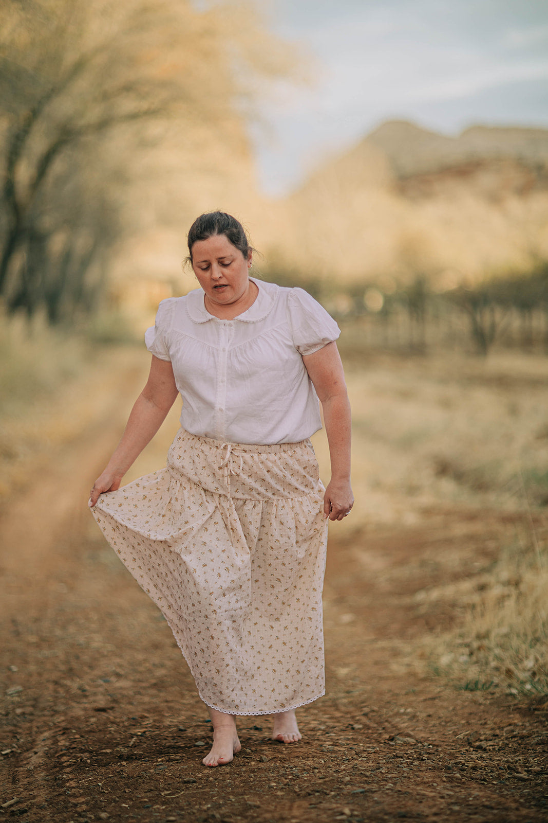 Model walks barefoot up a tree lined path. She is wearing a puff sleeved button down blouse. It is white with a gathered bodice and peter pan color.