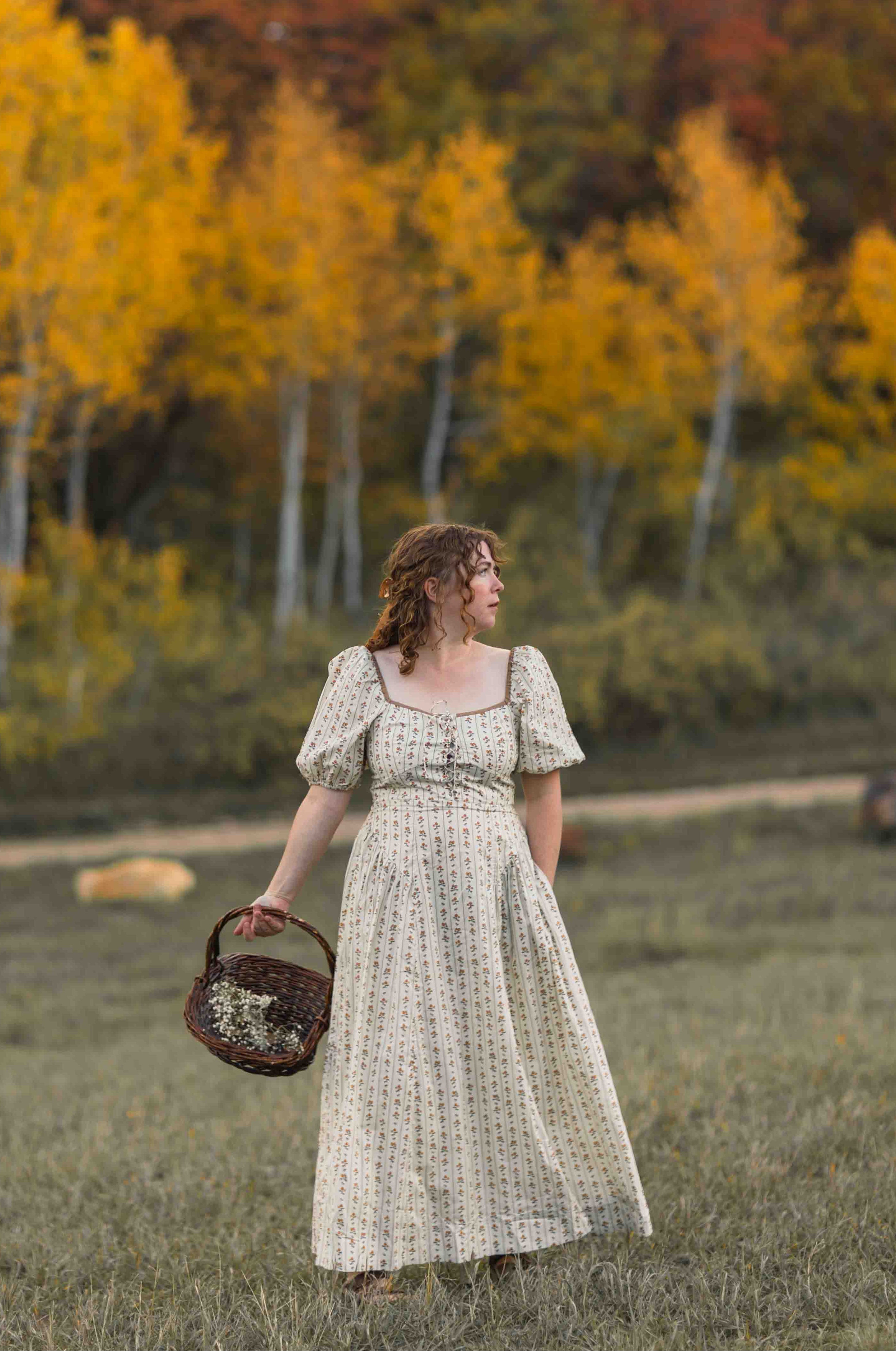A woman in a cream-colored, short-sleeved floral dress stands in a grassy field holding a woven basket, with a background of vibrant autumn trees in yellow, orange, and red.