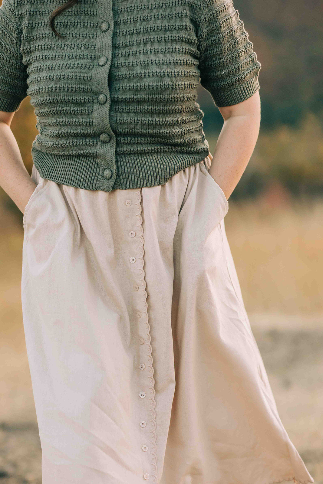 Person wearing a green knitted cardigan and beige button down skirt with a blurred natural background