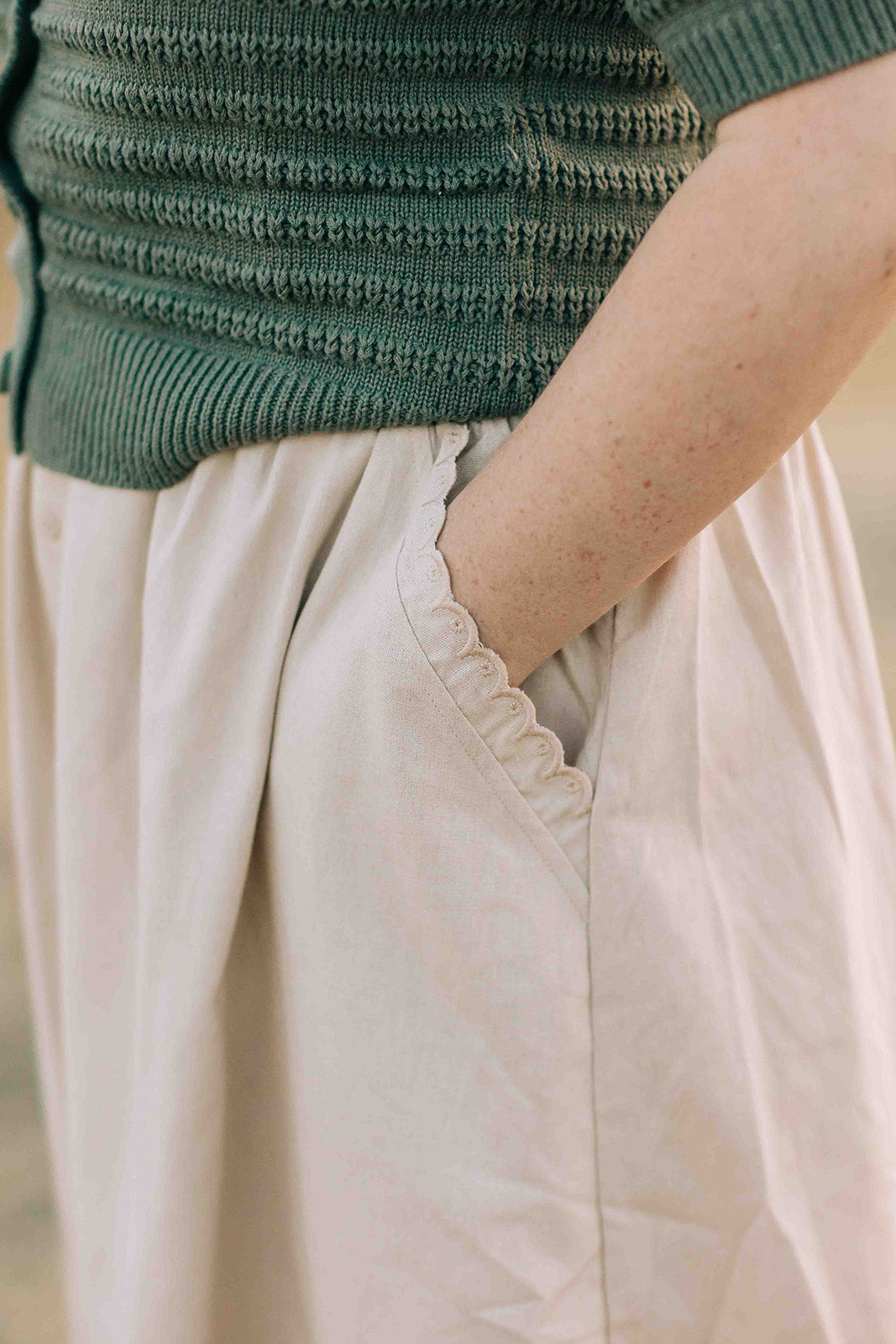 Person wearing a green sweater and beige scalloped trim skirt with a blurred background